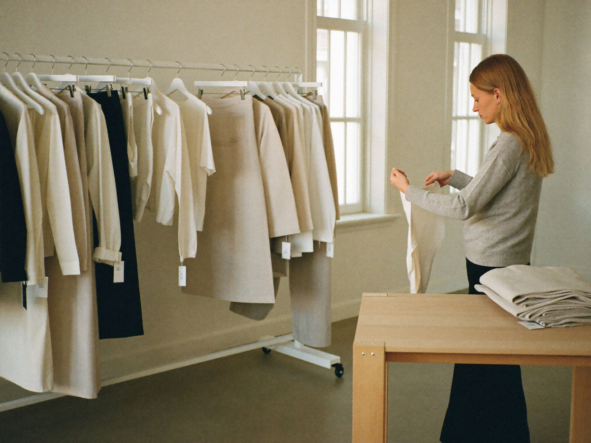 A bright studio with a rail of neutral womenswear and folded knitwear on a pale table